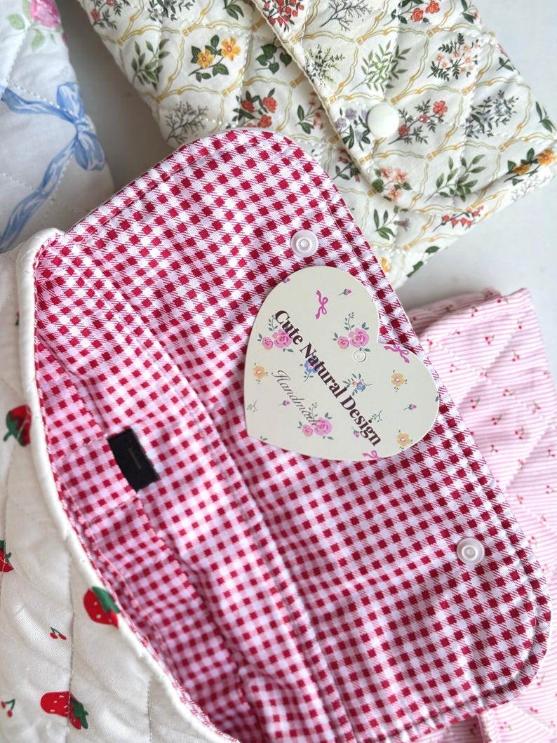 May include: Close-up of several quilted fabric pouches with different patterns. One features a red and white gingham lining, another has a floral design, and a third has a strawberry pattern. A heart-shaped tag reads "Cute Natural Design Handmade."