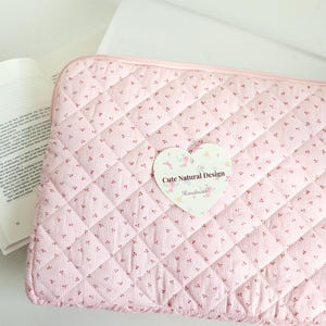 May include: Pink quilted laptop sleeve with a floral pattern and a heart-shaped tag that reads "Cute Natural Design". A book with text is open next to the sleeve. The sleeve is approximately 13 inches wide and 10 inches tall.