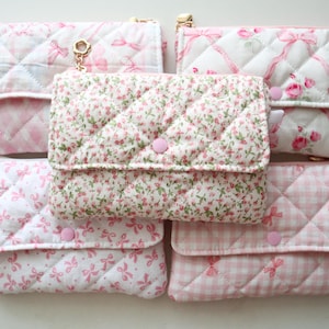 May include: Five rectangular quilted pouches with flap closures and pink snap buttons. The pouches feature various patterns including pink and white gingham, small floral prints, and pink ribbon motifs on white fabric, arranged in a grid formation.