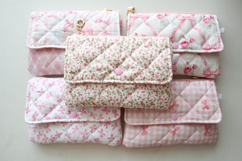 May include: Five rectangular quilted pouches with flap closures and pink snap buttons. The pouches feature various patterns including pink and white gingham, small floral prints, and pink ribbon motifs on white fabric, arranged in a grid formation.