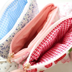 May include: Three quilted zippered pouches with distinct interior linings. One features a blue gingham interior, another a solid light pink lining, and the third a red and white gingham interior. Each pouch has a gold-toned metal zipper pull.