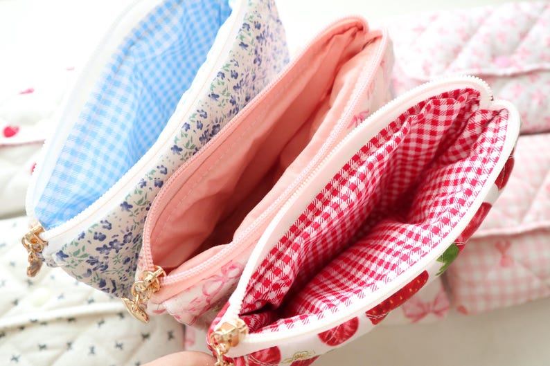 May include: Three quilted zippered pouches with distinct interior linings. One features a blue gingham interior, another a solid light pink lining, and the third a red and white gingham interior. Each pouch has a gold-toned metal zipper pull.