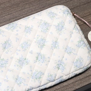 May include: Cream-colored quilted laptop sleeve with a floral pattern in light blue. The sleeve has a zipper closure and a heart-shaped tag that reads "Cute Natural Design Handmade".
