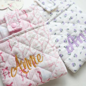 Può includere: Due pochette in tessuto trapuntato con chiusure a pressione. La pochette a sinistra è rosa con un motivo a nastri e il nome Annie ricamato in oro. La pochette a destra è bianca con un motivo floreale viola e il nome Ann ricamato in viola.
