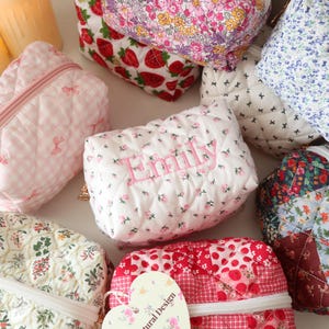 May include: A collection of quilted fabric pouches in various patterns including strawberries, floral prints, and gingham checks. One white pouch features the name Emily embroidered in pink thread. Each pouch has a zipper closure and soft texture.