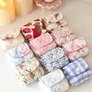 Pu&ograve; includere: Una collezione di piccole pochette in tessuto trapuntato con vari motivi, tra cui fragole, stampe floreali e quadretti vichy blu. Ogni pochette quadrata &egrave; dotata di una patta chiusa da un bottone a pressione in plastica.