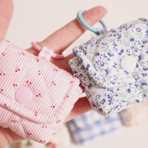 Pu&ograve; includere: Due piccole pochette in tessuto trapuntato con chiusure a scatto e portachiavi. Una pochette presenta un motivo a righe rosa e bianche con minuscole ciliegie rosse, mentre l'altra mostra una stampa floreale blu e bianca. Texture morbida e imbottita.