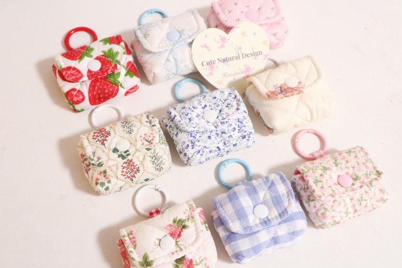Pu&ograve; includere: Una collezione di piccole pochette in tessuto trapuntato con vari motivi, tra cui fragole, stampe floreali e quadretti vichy blu. Ogni pochette &egrave; dotata di chiusura a bottone automatico e di un anello circolare in plastica per appenderla.