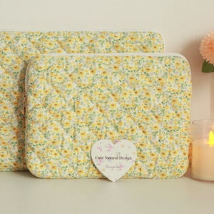 May include: Two quilted laptop sleeves with a yellow floral pattern. The sleeves have a white zipper and a diamond-shaped stitching pattern. A heart-shaped tag with the text "Cute Natural Design" is in front of the sleeves.