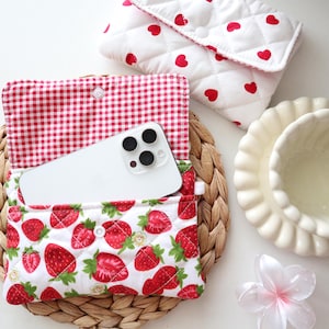 May include: A quilted fabric pouch featuring a red strawberry print on a white background with a red and white gingham interior lining, containing a white smartphone. A second quilted pouch with red hearts is visible nearby.