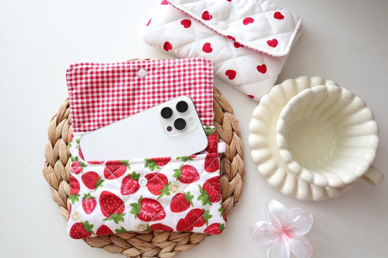 May include: A quilted fabric pouch featuring a red strawberry print on a white background with a red and white gingham interior lining, containing a white smartphone. A second quilted pouch with red hearts is visible nearby.