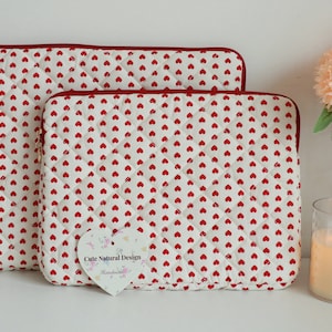 May include: Two quilted laptop sleeves with a white background and a pattern of red hearts. The edges are trimmed with a dark red fabric. A small tag with the text "Cute Natural Design" is attached to the front.
