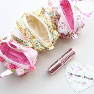 May include: Three small fabric pouches with handles, featuring floral and gingham patterns in pink, yellow, and white. A metallic lipstick tube and a heart-shaped card with the text "Cute Natural Design" and "Handmade" are placed nearby.