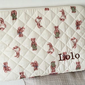May include: Cream-colored quilted laptop sleeve with a repeating pattern of teddy bears in various poses. The bears are in shades of brown and wear outfits. The name "Lolo" is embroidered in brown on the lower right. A gold zipper is visible.