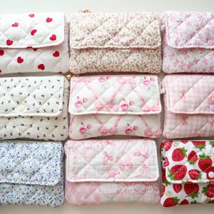 May include: A collection of nine rectangular quilted pouches arranged in a grid. Each pouch features a fold-over flap with a snap closure and various patterns including red hearts, floral prints, pink bows, gingham, and strawberries on white fabric.