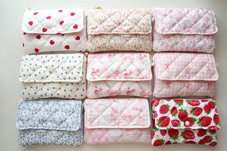 May include: A collection of nine rectangular quilted pouches arranged in a grid. Each pouch features a fold-over flap with a snap closure and various patterns including red hearts, floral prints, pink bows, gingham, and strawberries on white fabric.