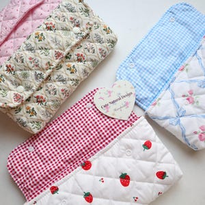May include: A collection of quilted fabric pouches in various patterns. Designs include pink stripes, floral, gingham, and strawberry prints. Each pouch has a snap closure. A heart-shaped tag reads "Cute Natural Design."
