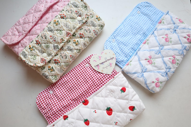 May include: A collection of quilted fabric pouches in various patterns. Designs include pink stripes, floral, gingham, and strawberry prints. Each pouch has a snap closure. A heart-shaped tag reads "Cute Natural Design."