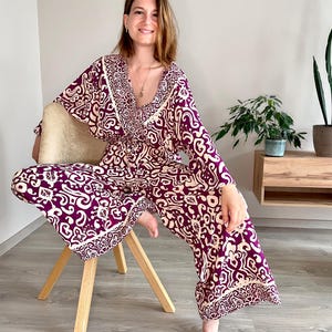 Könnte beinhalten: Eine Frau trägt einen lila- und cremefarbenen Jumpsuit mit weiten Beinen. Der Jumpsuit hat einen V-Ausschnitt und lange Ärmel. Die Frau sitzt auf einem Holzstuhl.