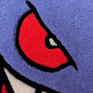 May include: Close-up of a blue, red, black, and white rug with a cartoon monster eye design. The eye is red with a black outline and a white highlight. The rug has a textured surface.