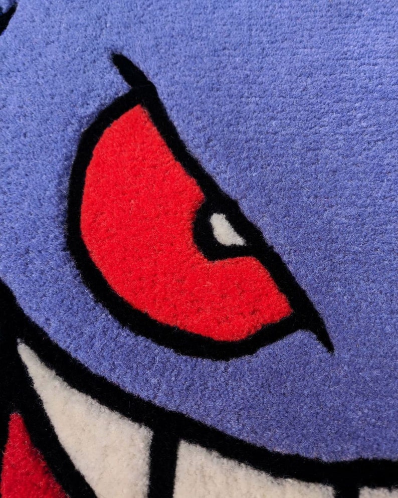 May include: Close-up of a blue, red, black, and white rug with a cartoon monster eye design. The eye is red with a black outline and a white highlight. The rug has a textured surface.