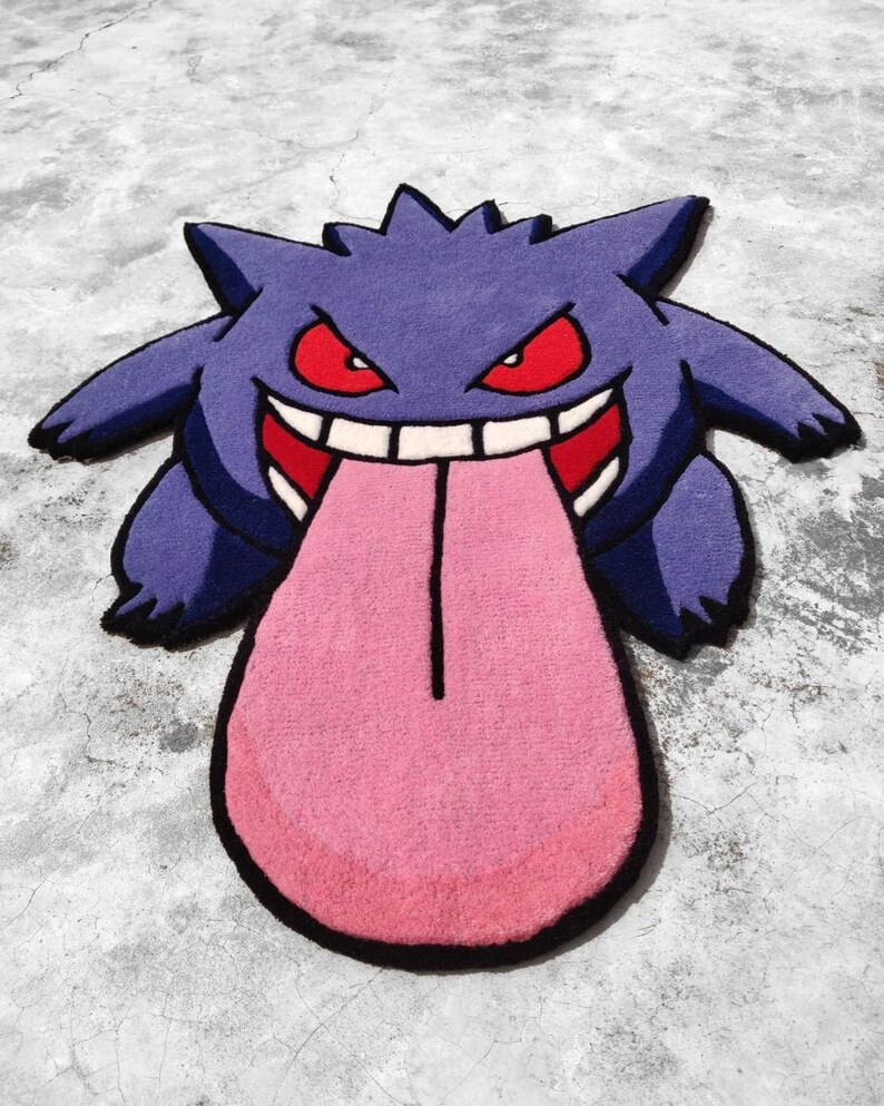 May include: A purple and black rug shaped like a cartoon character with a large pink tongue. The character has red eyes, a wide grin, and pointed ears. The rug is placed on a gray concrete surface.