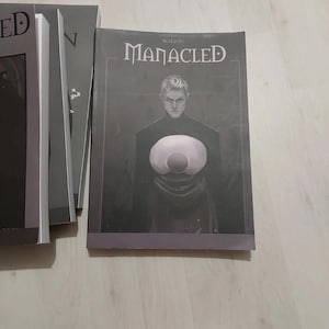Op de afbeelding: Stapel boeken met de titel "MANACLED" op de omslag. De omslag toont een persoon met een groot, cirkelvormig object. De boeken liggen op een lichtgekleurd houten oppervlak.