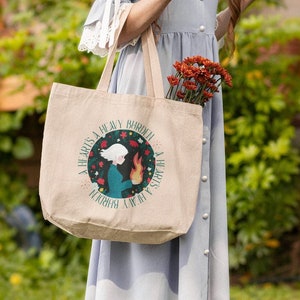 Puede incluir: Una bolsa de tela beige con un diseño floral y el texto "Hearts are a heavy burden, a heavy burden to bear." La bolsa la lleva una persona que lleva un vestido azul claro.