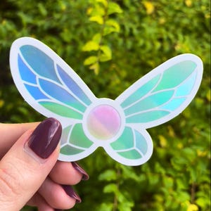 May include: A holographic iridescent butterfly sticker with a pink and blue center. The sticker is on a white background.