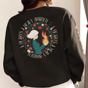 May include: Black sweatshirt with a circular design reading "A Heart's A Heavy Burden." The graphic shows a person holding a flame, encircled by flowers. The sleeve has floral details.