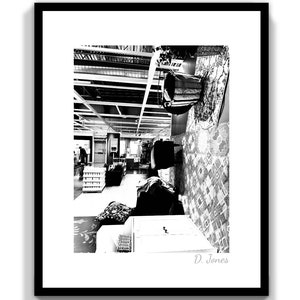 May include: Black and white photograph of a retail store interior. The image shows a wide view of the store, with various displays, furniture, and decorative items. The store has a high ceiling with exposed beams and lighting fixtures. A patterned rug hangs on the wall.