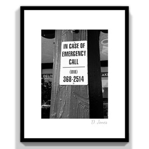 May include: Black and white framed print featuring a sign on a wooden post. The sign reads "IN CASE OF EMERGENCY CALL" followed by a phone number (818) 368-2514. The print is signed "D. Jones" in the bottom right corner.