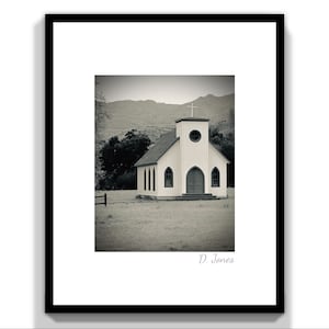 May include: A black and white photograph of a small, white church with a cross on the roof. The church is surrounded by grass and trees, and there are mountains in the background.