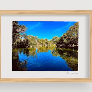 Puede incluir: Una fotografía enmarcada de un lago rodeado de árboles con un cielo azul y nubes blancas. El agua refleja el cielo y los árboles. La firma "D. Jones" está en la esquina inferior derecha de la imagen.