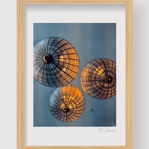 Puede incluir: Tres luminarias redondas tejidas cuelgan de un techo contra un cielo azul. Las luminarias están iluminadas desde adentro, proyectando un brillo cálido. La imagen está firmada "D. Jones" en la esquina inferior derecha.