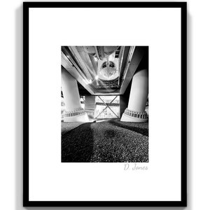 Può includere: Fotografia in bianco e nero della parte inferiore di un ponte. L'immagine mostra i supporti in cemento del ponte e le travi in acciaio che li collegano. La foto è stata scattata da un angolo basso, guardando verso l'alto il ponte.