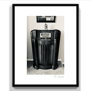 May include: A vintage, black wooden radio with a silver face and a grid-like speaker. The radio is sitting on a carpeted floor in a room with a framed picture on the wall behind it.
