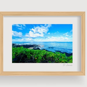 May include: A framed photograph of a seascape with a blue sky, white clouds, and a rocky coastline. The ocean is a deep blue color and the rocks are black. The photo is framed in a light wood frame. The photographer's signature, "D. Jones", is in the bottom right corner.