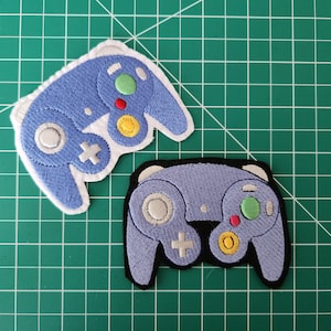 May include: Two blue embroidered patches of a Nintendo GameCube controller. The patches are on a green cutting mat.