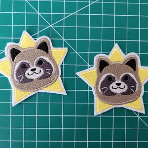 May include: Two embroidered iron-on patches featuring a cartoon raccoon face inside a yellow star. The raccoon has a brown face, black eyes, and a white nose.
