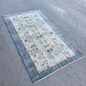 May include: Rectangular rug with a floral pattern in shades of beige, purple, and green. The rug has a faded appearance with a blue border. The rug is placed on a gray surface.
