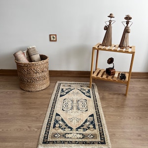 May include: A rectangular rug with a beige central field and a dark blue border. The rug features a symmetrical geometric pattern with floral and stylized bird motifs. A woven basket with rolled textiles is in the background.