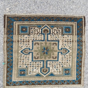 May include: A small, square rug with a geometric design. The rug features a central cross-like pattern in shades of beige, blue, and green, framed by a border of brown and blue. The rug's dimensions are approximately 24 inches by 24 inches.