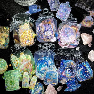 以下が含まれることがあります： さまざまなデザインの瓶、ボトル、ティーカップをあしらった装飾ステッカーのコレクション。蝶が入った瓶もあれば、天体のシーンが入った瓶もあります。ステッカーはホログラフィックで虹色の仕上がりで、白いアウトラインと暗い背景があります。