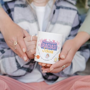 Könnte beinhalten: Eine weiße Keramiktasse mit einem rosa und violetten Design. Die Tasse trägt den Text "Muchas Gracias! Jehova" und eine kleine Illustration einer Person, die ein Herz hält.