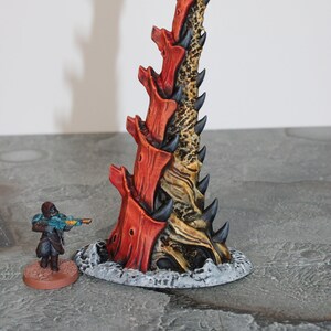 May include: A detailed miniature fantasy terrain piece, featuring a tall, spiky structure with red, brown, and black accents. A miniature figure with a rifle stands nearby, providing scale. The base is a textured, grey surface.