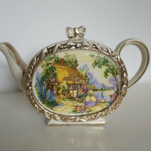 May include: A ceramic teapot featuring a central oval panel with a colourful illustration of a thatched-roof cottage, a garden, and a figure in a yellow dress. The teapot has a cream-coloured body with gold trim, a bow-shaped lid handle, and a spout.