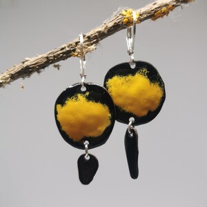 Puede incluir: Pendientes hechos a mano con un diseño negro y amarillo. Los pendientes son redondos, con un centro amarillo y un borde negro. De cada pendiente cuelga un pequeño dije en forma de lágrima de color marrón oscuro. Cuelgan de cierres de palanca plateados.