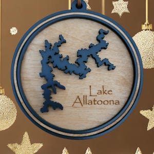 May include: Wooden Lake Allatoona ornament. The ornament features a detailed map of the lake in dark blue, set against a light wood grain background. The text "Lake Allatoona" is engraved below the map. The ornament is round with a dark blue border.