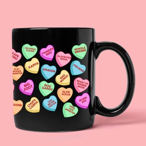 May include: Black ceramic mug decorated with colorful heart-shaped candies. Each candy has a different cryptid name, including "Bigfoot," "Kraken," and "Chupa Cabra." The mug has a curved handle and a pink background.
